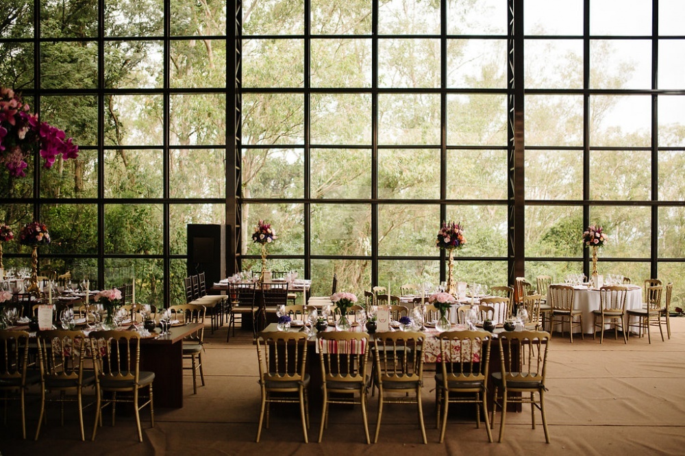 Mesas decoradas com arranjos florais e cadeiras douradas com grandes janelas de vidro